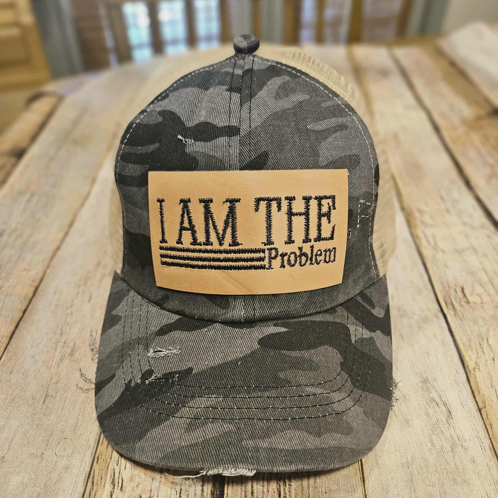 Camouflage cap with 'I AM THE Problem' patch on a wooden surface